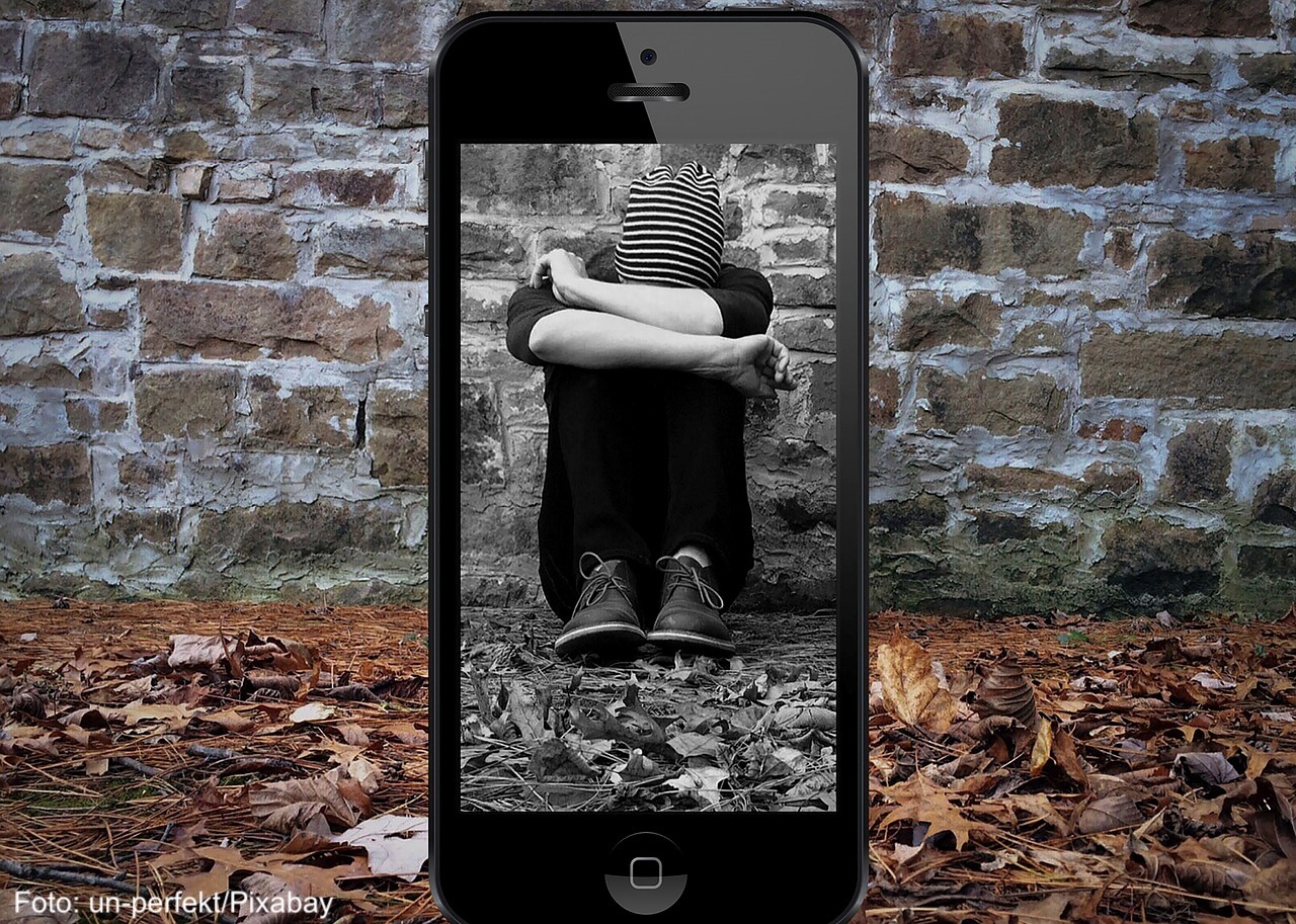 Das Foto zeigt ein Smartphone, das ein Bild von einem Jugendlichen macht. Der Jugendliche sitzt auf dem Boden und versteckt sein Gesicht.