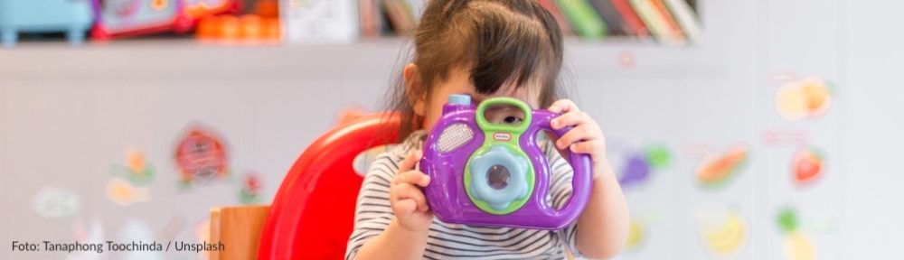 Auf dem Bild ist ein kleines Mädchen zu sehen, dass durch eine digitale Kinderkamera schaut.